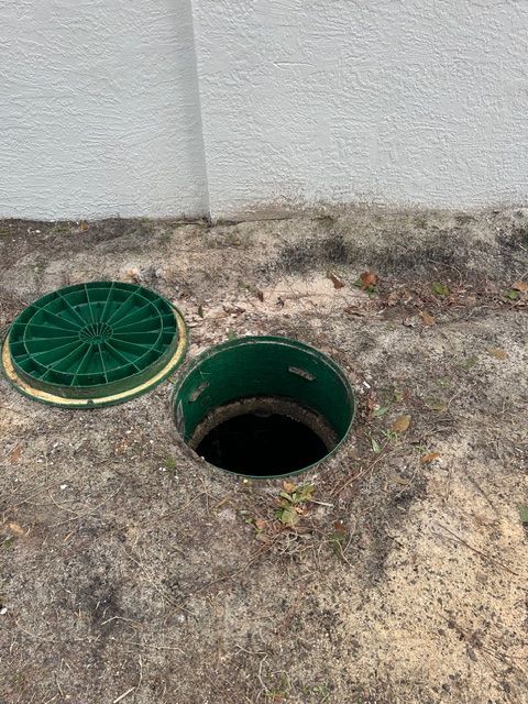 Open green septic tank access in the ground beside a white wall, with its lid nearby.