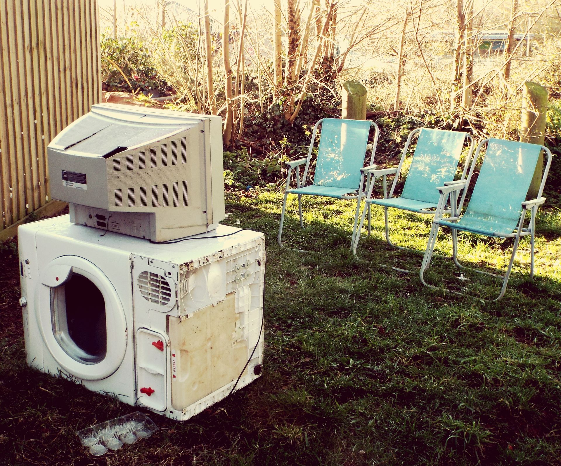 An old television sits atop a dryer on grass, with three blue lawn chairs in the background.