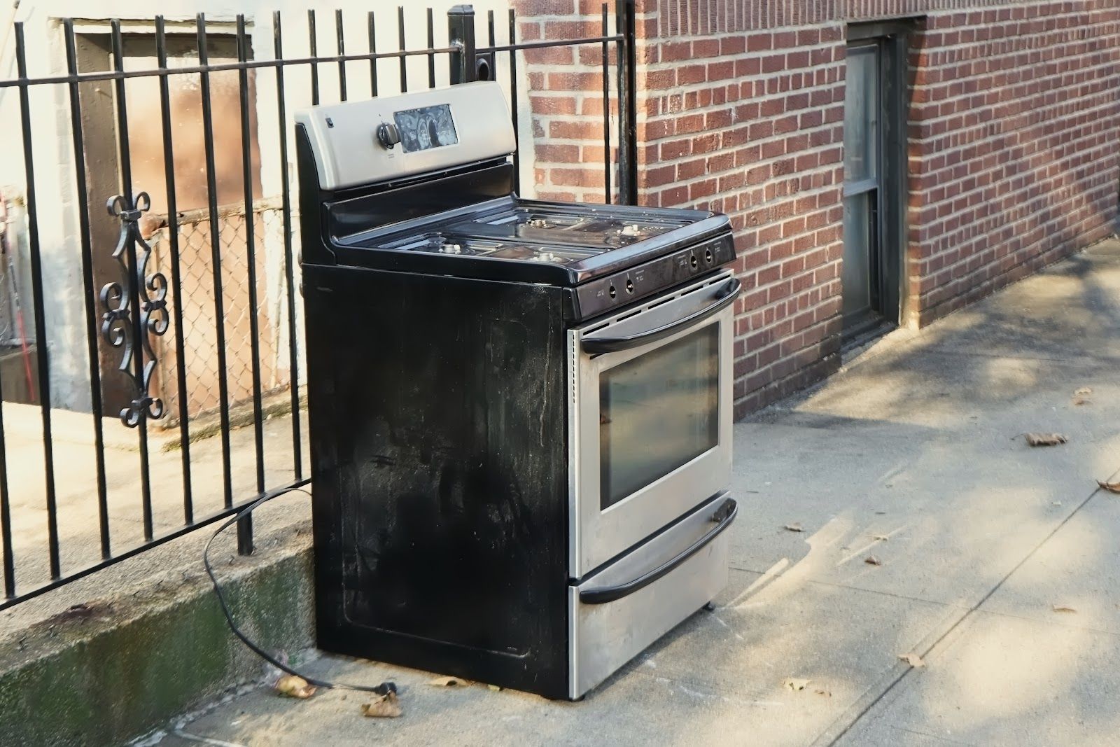 Black and silver oven on a sidewalk next to a brick building and a metal fence.