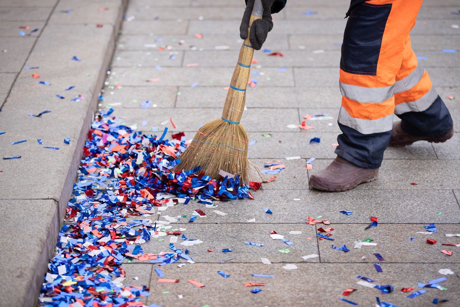 Worker in orange and blue overalls sweeping confetti from a sidewalk with a straw broom.