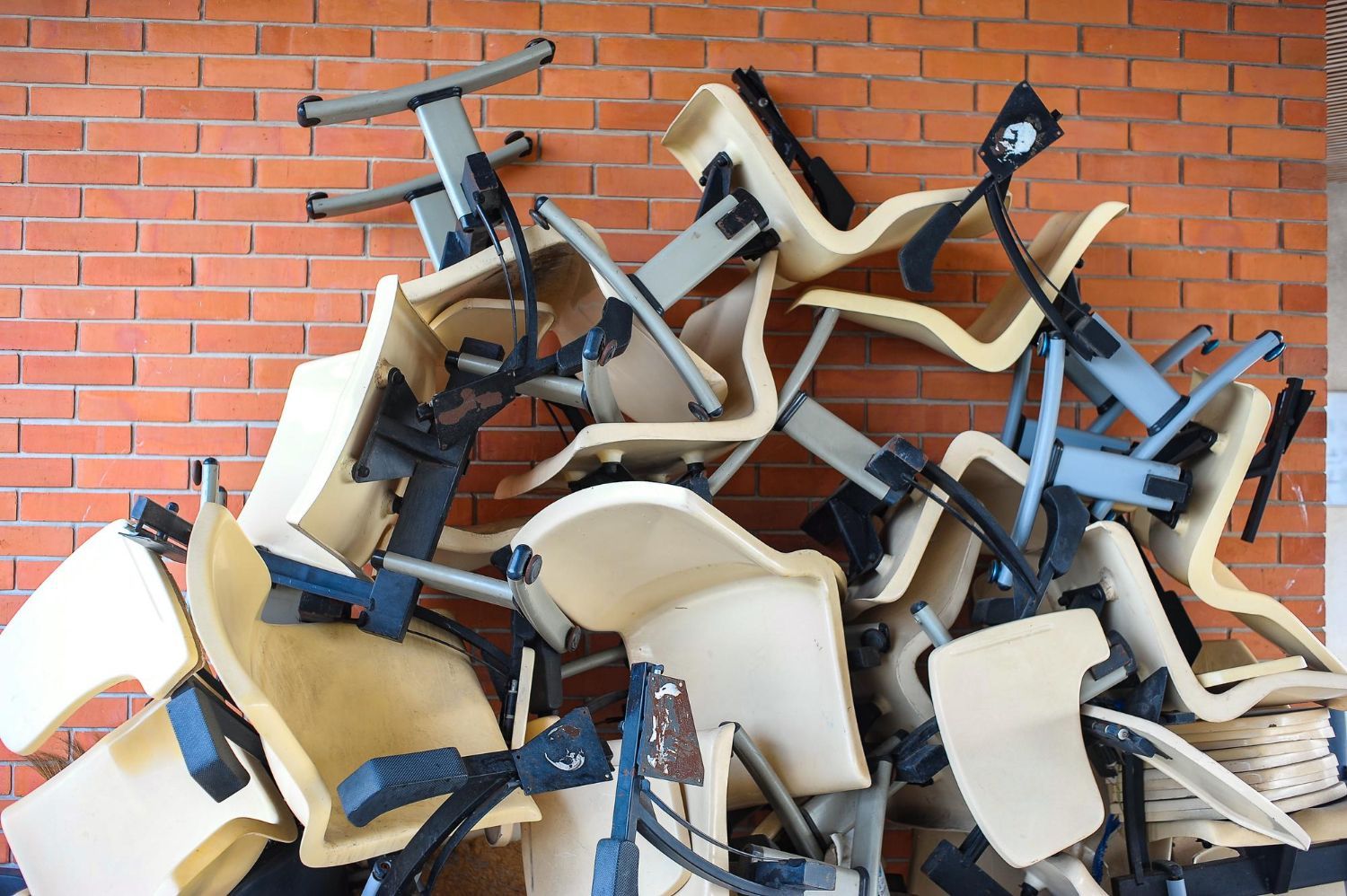 Pile of beige and black office chairs against a red brick wall. Chairs are stacked haphazardly.