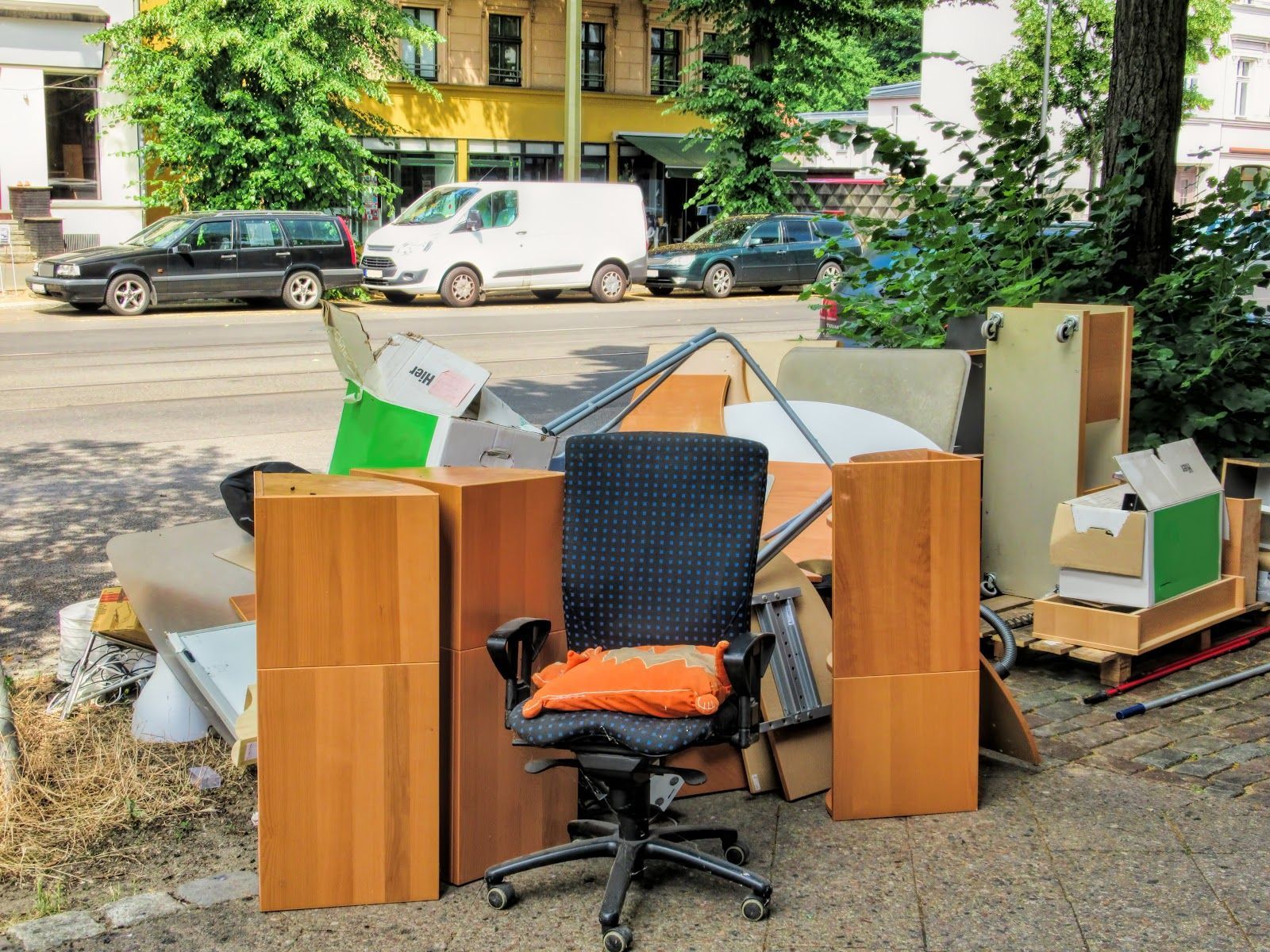 Pile of discarded furniture and cardboard on a city street; cars and buildings in background.