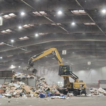 A yellow excavator is moving a pile of cardboard in a large warehouse.