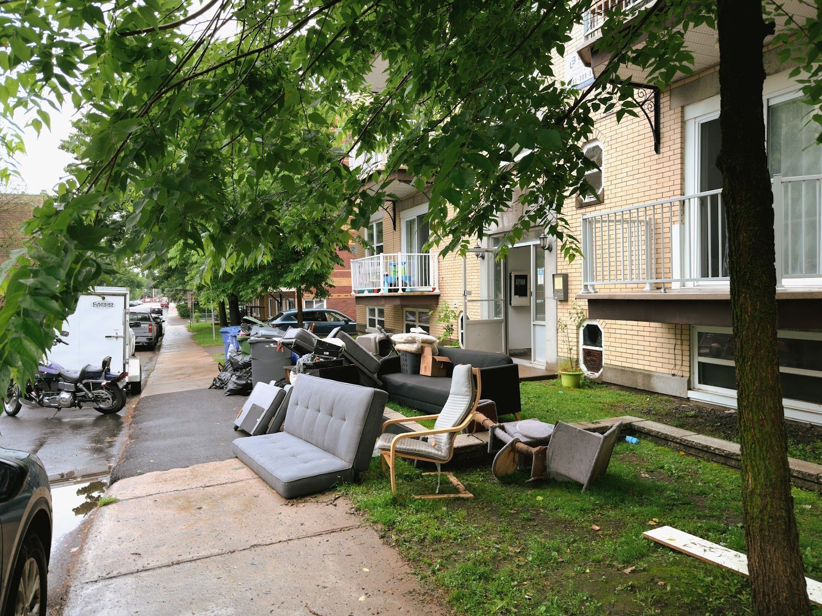 Street scene: discarded furniture piled on grass and sidewalk in front of a brick apartment building.
