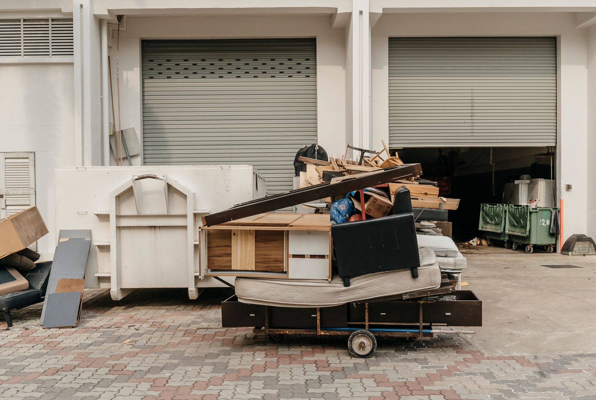 A person loads discarded furniture onto a cart near a dumpster in an industrial setting.