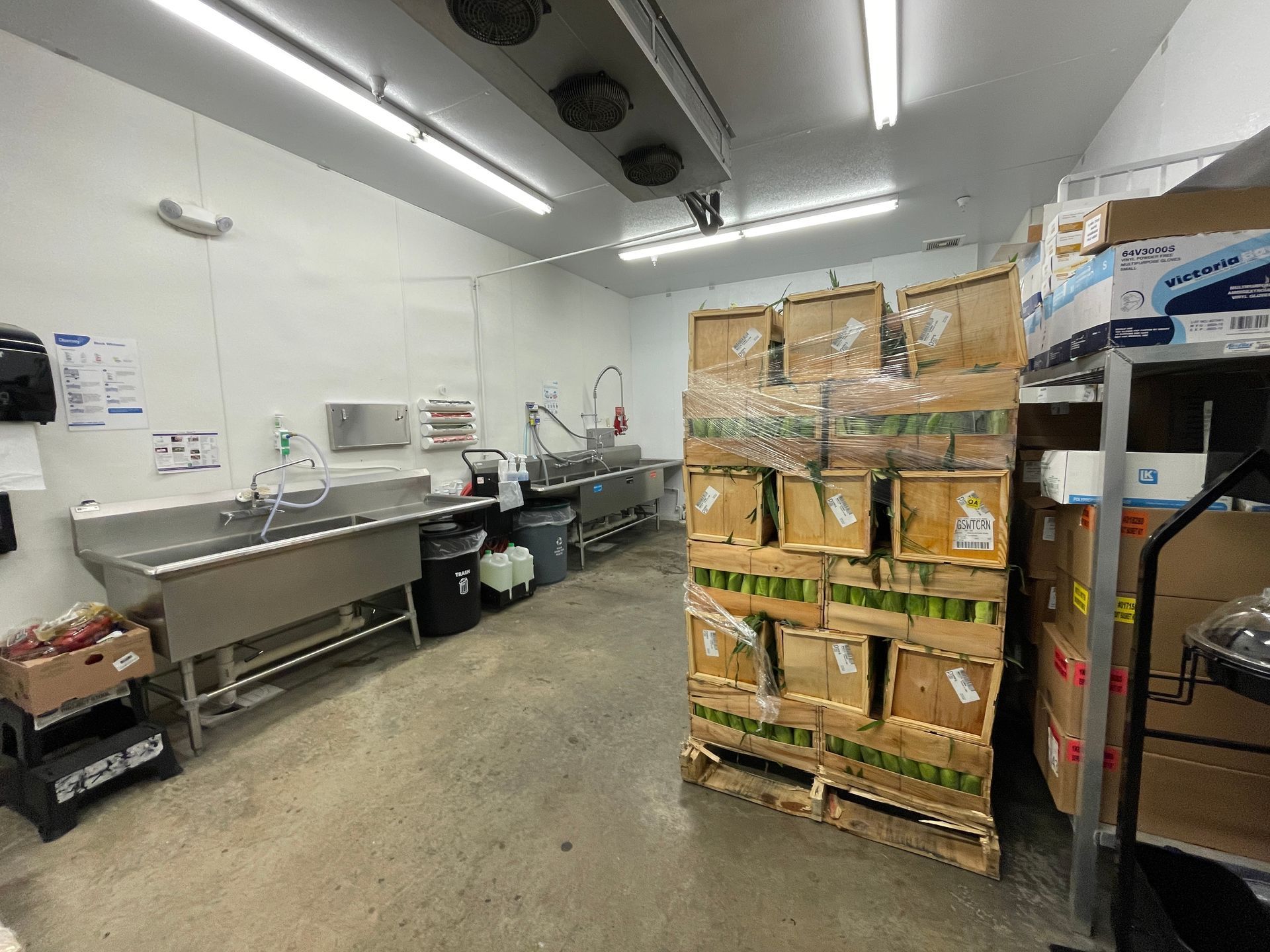 A commercial kitchen with stainless steel sinks, a pallet of boxes, and bright overhead lights.
