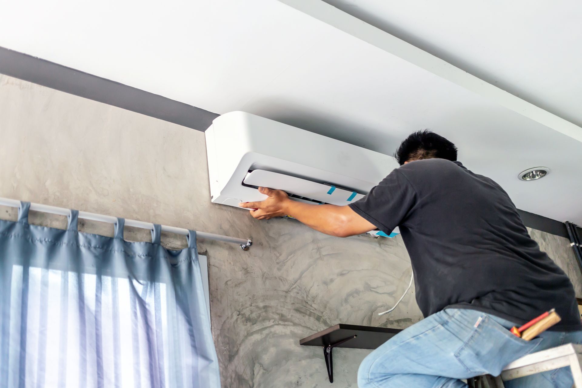 A man is sitting on a ladder installing an air conditioner.