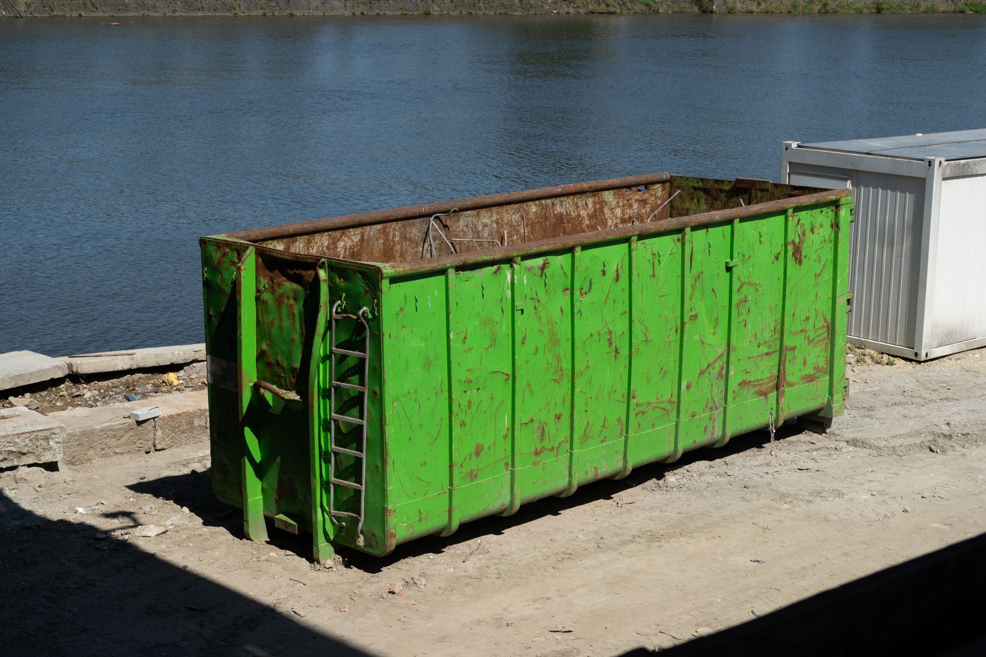 A green dumpster is sitting on the ground next to a body of water.