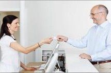 smiling lady standing behind a waist height desk giving a card to a smiling gentleman.