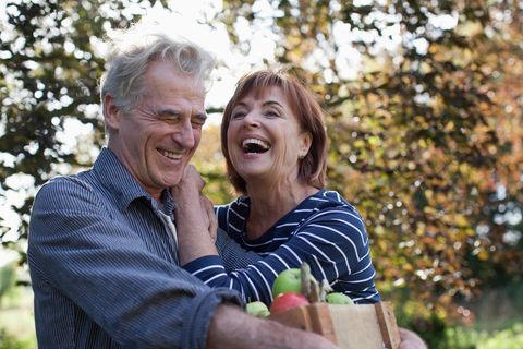 mature couple laughing in an orchard. Man is holding a small crate of apples.