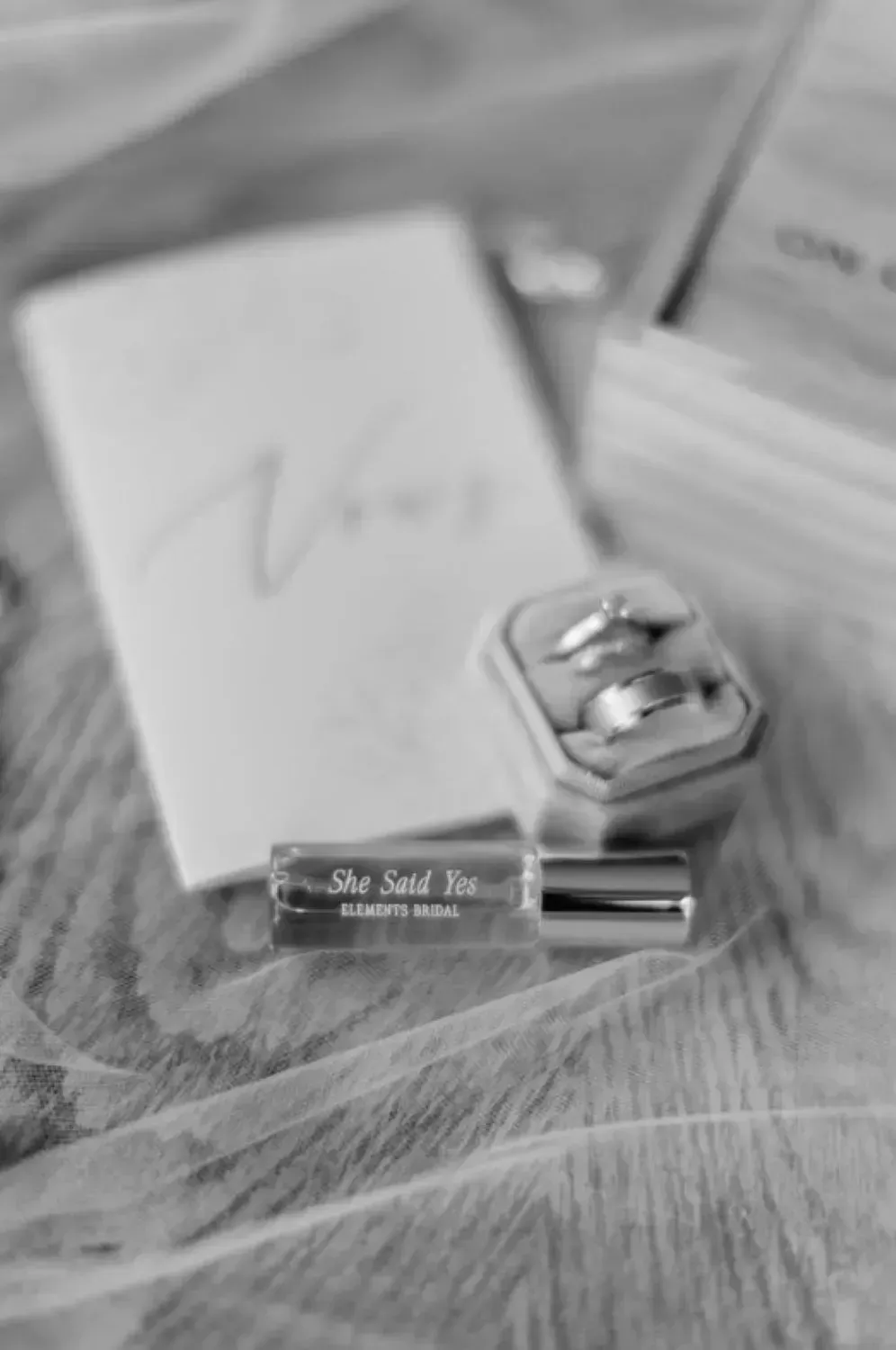 A black and white photo of a wedding ring and a perfume bottle on a table.