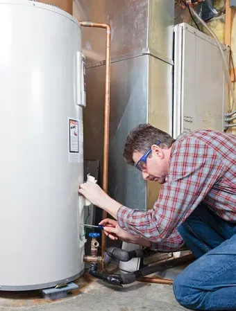 Man in plaid shirt and safety glasses inspecting a water heater in a utility room.