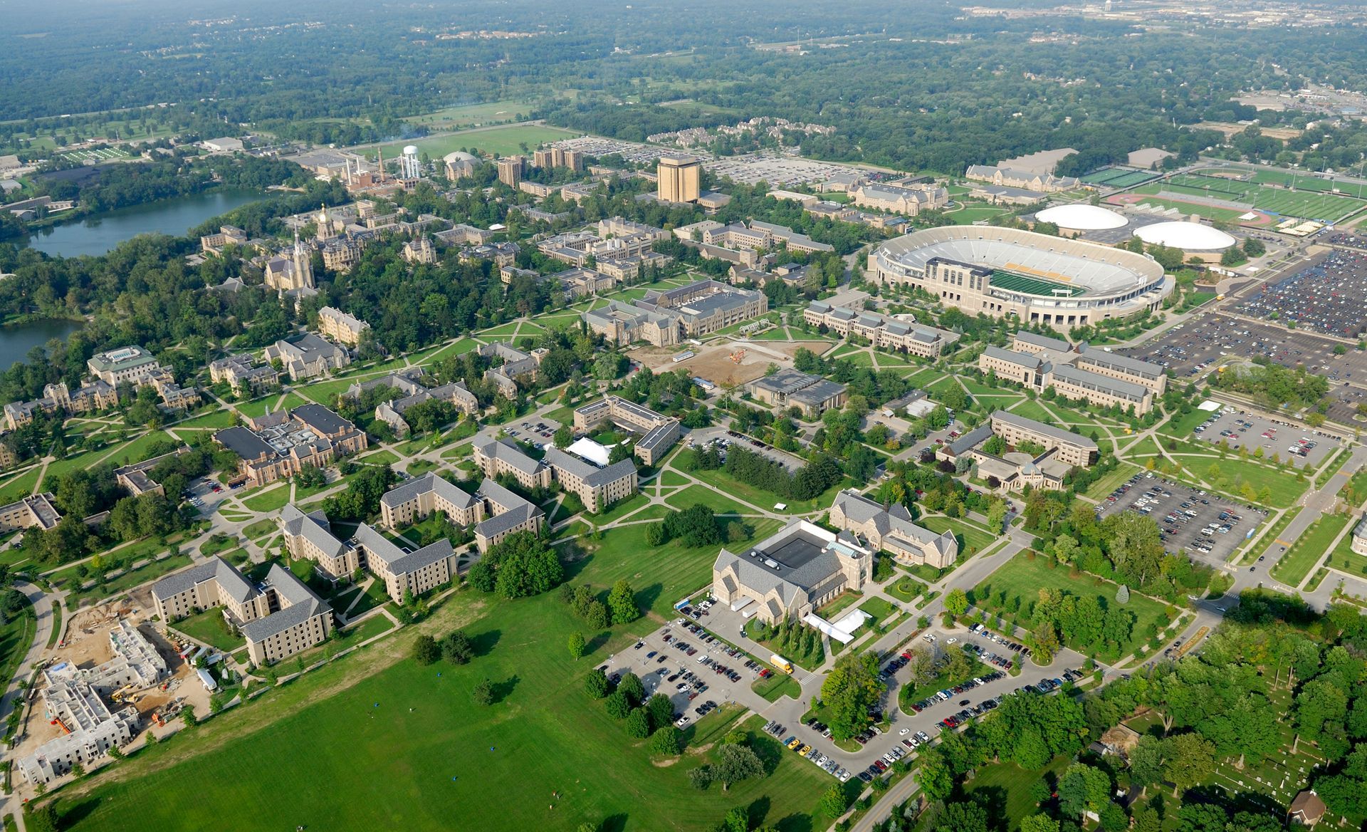 Handshake between a person in a blue jumpsuit holding a toolbox and a person wearing jeans and a light-colored top, by a doorway. Aerial view of a large university campus with buildings, sports stadium, and green spaces.