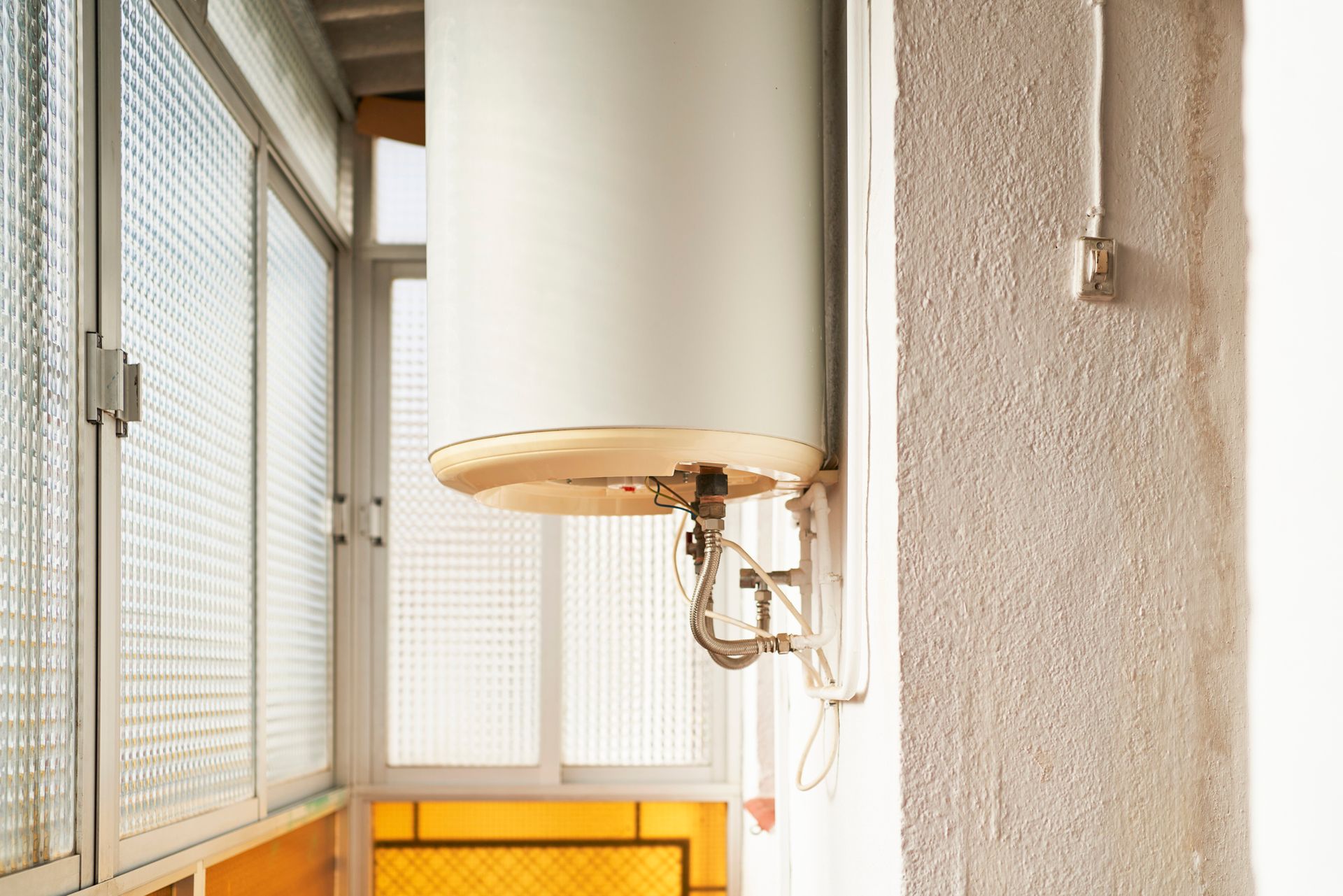 Water heater, white, mounted on a textured wall, near a windowed balcony.