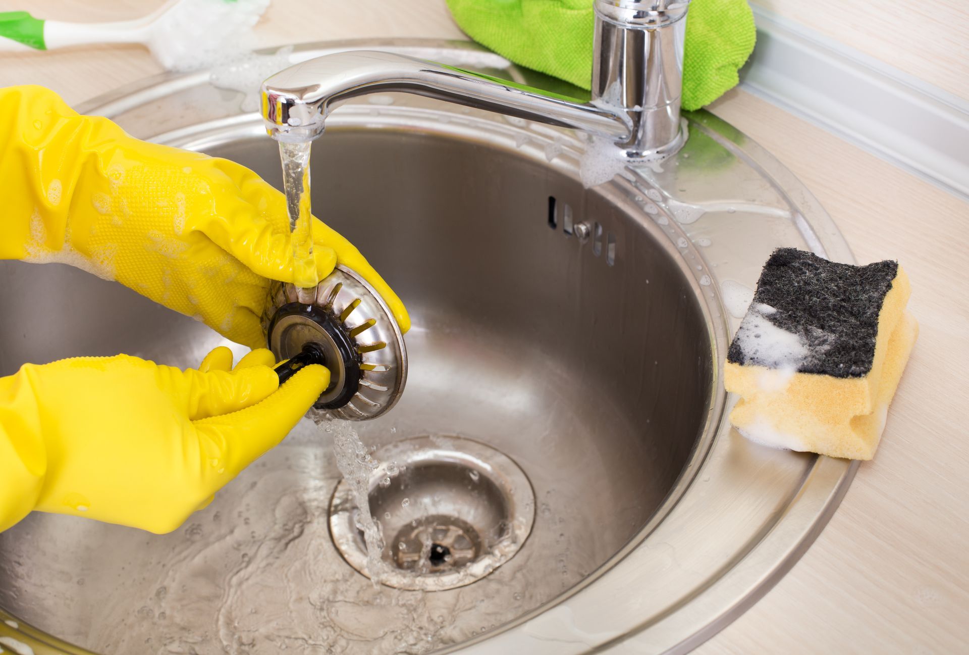 Hands in yellow gloves cleaning a kitchen sink strainer under running water.