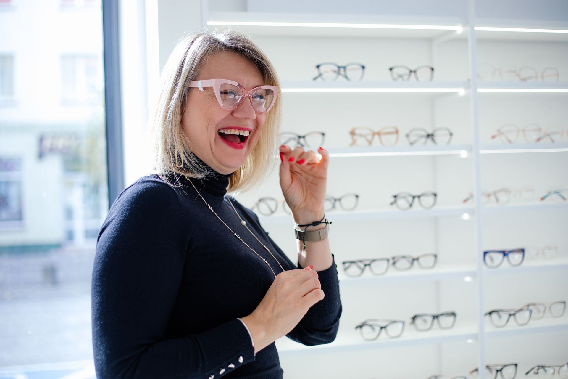 Happy blonde woman trying on glasses in a brightly lit optician store, showcasing a wide selection o