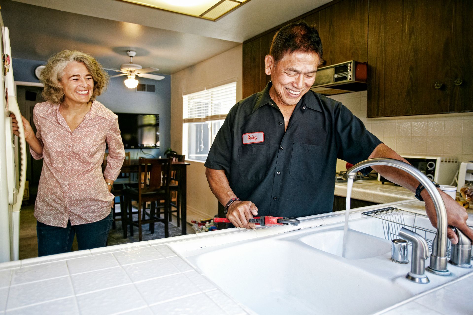 A professional plumber is checking a kitchen sink faucet with the customer next to him.