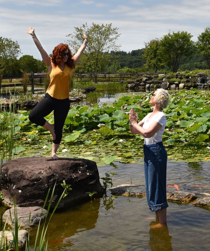 Cassandra Storm in tree pose on a rock and Liz Tattersall holding hands in prayer standing in a lily pond