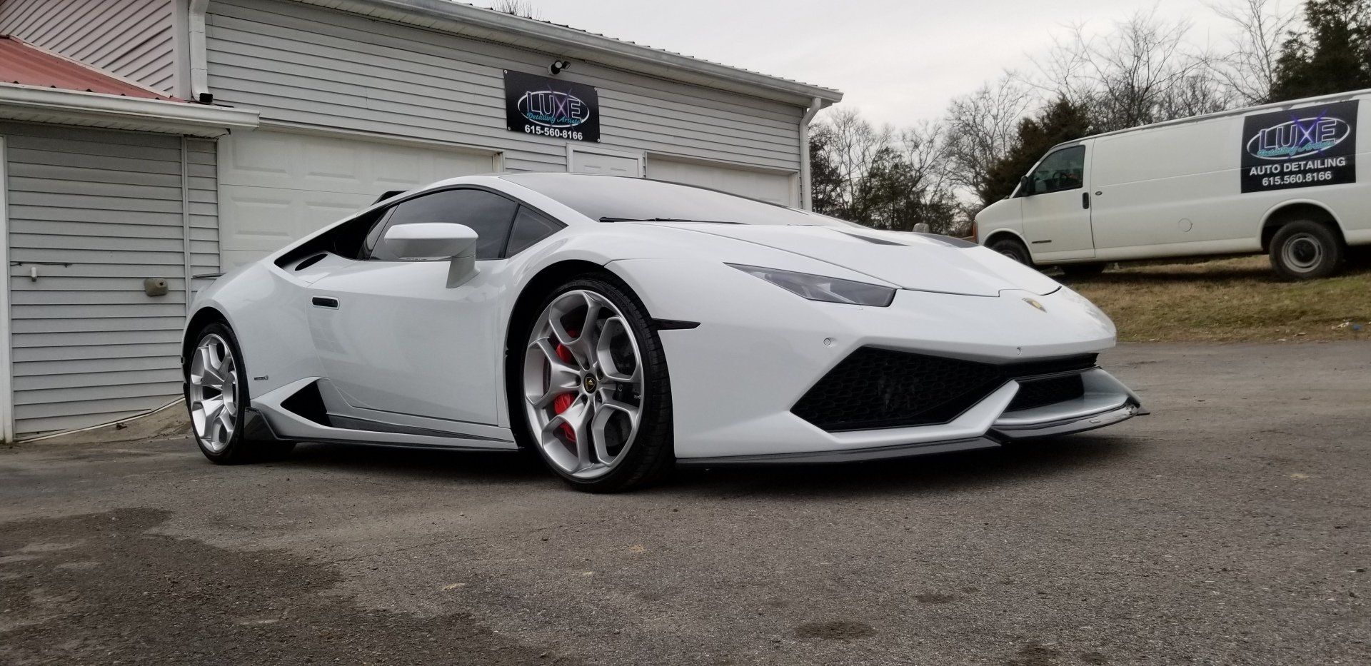 A white Lamborghini sports car parked in front of a building with a white van in the background.