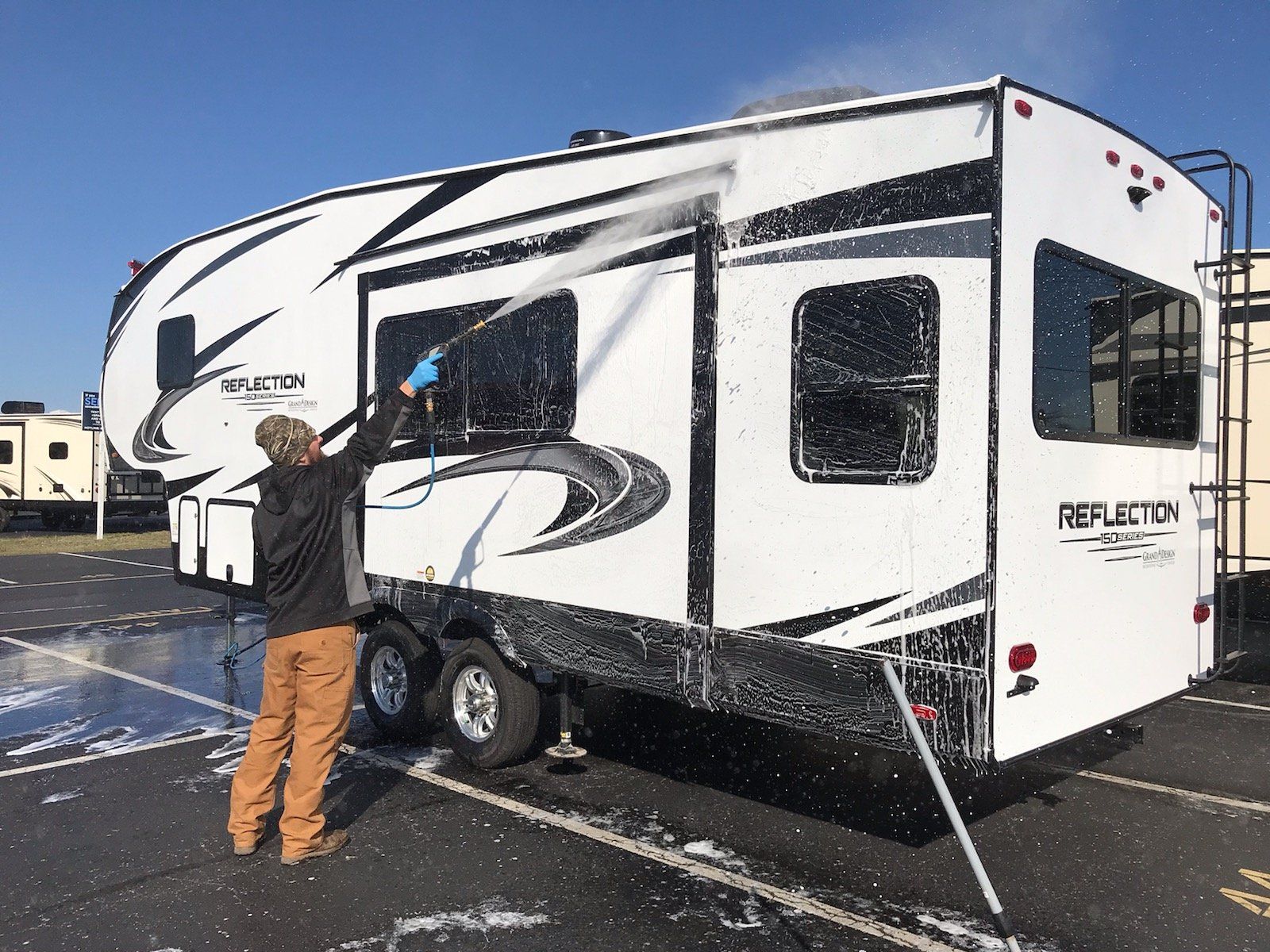 Person washing a white and black Reflection RV with a pressure washer outdoors.