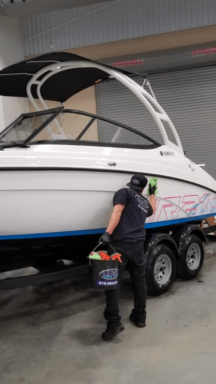 A man cleaning a white boat with blue accents in a garage, holding a bucket of cleaning supplies.