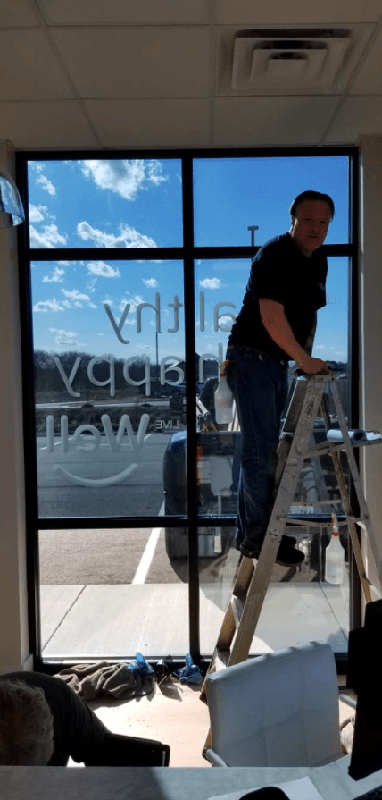 Man on ladder installing window graphics. Sky visible through window.