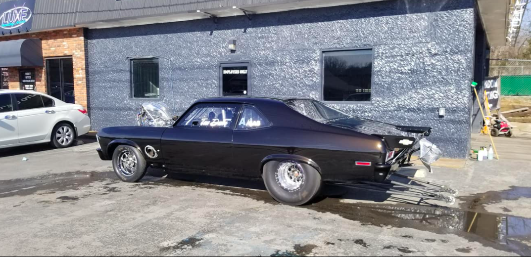 Dark muscle car parked in front of a building. Gray concrete ground with water and a white car in the background.
