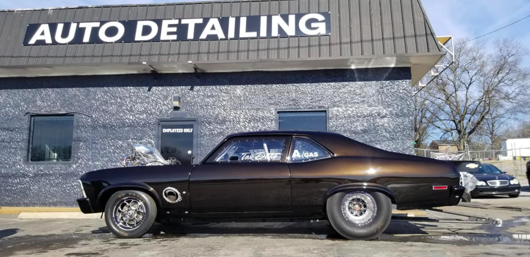 A dark brown classic car in front of an auto detailing shop.