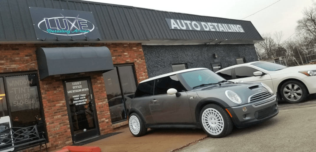 A gray Mini Cooper parked in front of an auto detailing shop, a white car is behind it.