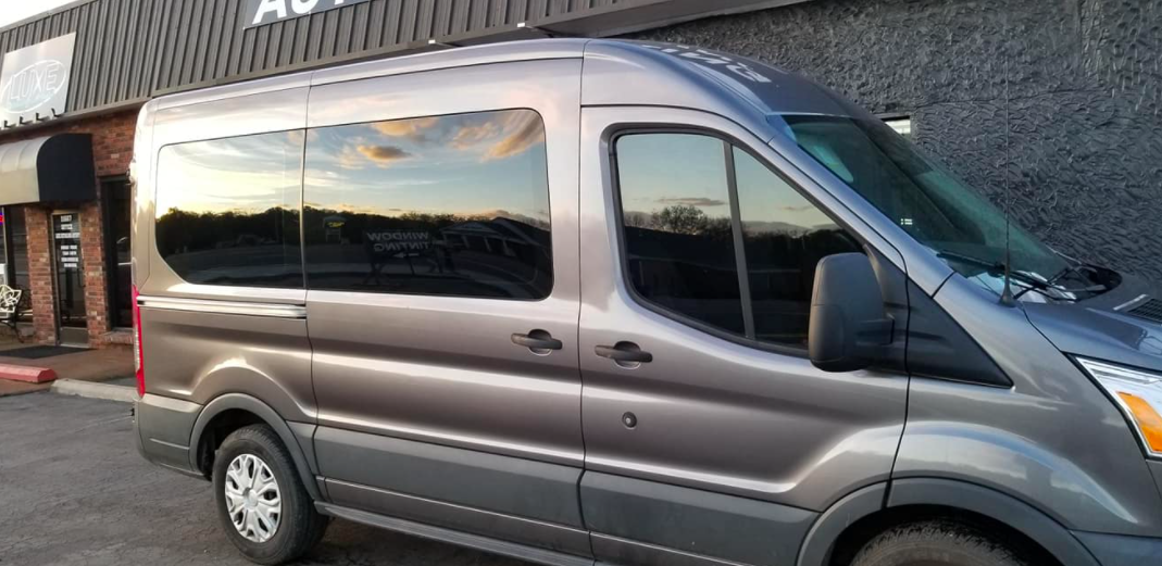 A grey passenger van parked in front of a building with tinted windows reflecting the sky.