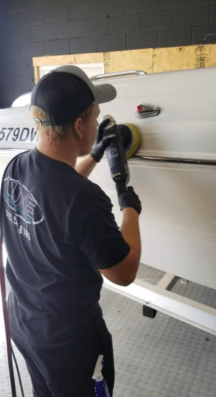 Person polishing a white boat with a power buffer. They wear a hat, gloves, and black shirt.