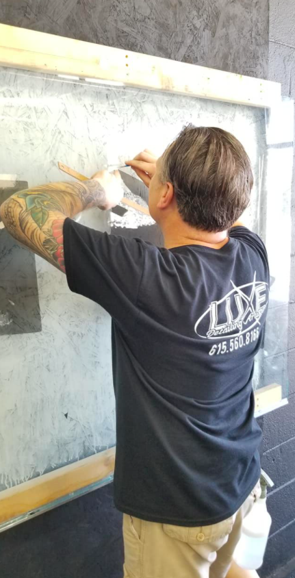 Man with tattoos works on artwork in front of a window; he's wearing a black shirt and tan pants.