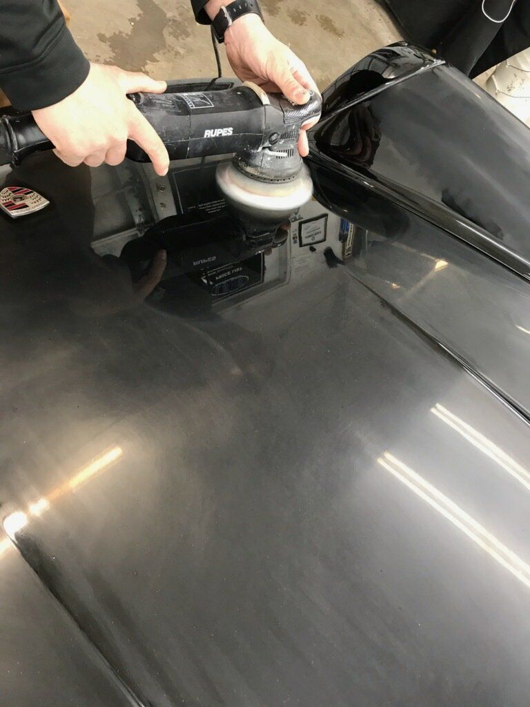 Person polishing a shiny black car hood with a buffer tool.