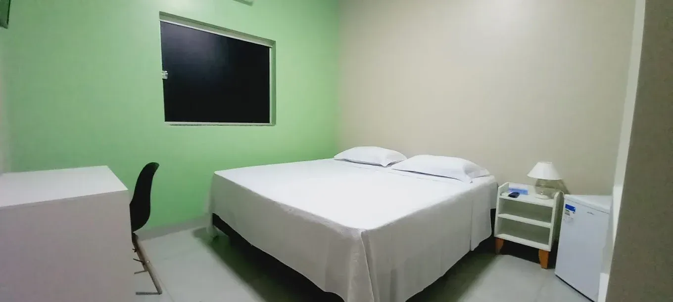 Um quarto com cama, mesa de cabeceira, geladeira e janela.