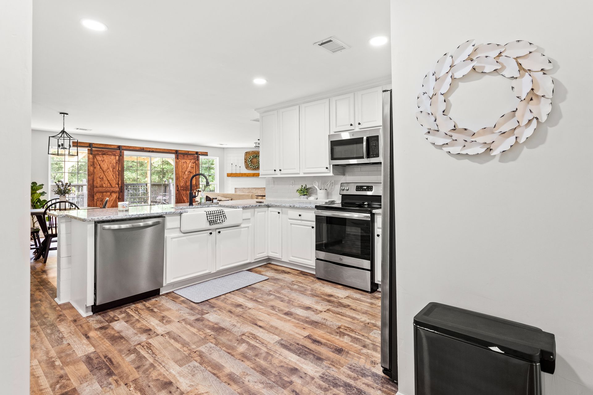 A kitchen with white cabinets , stainless steel appliances , wooden floors and a wreath on the wall.