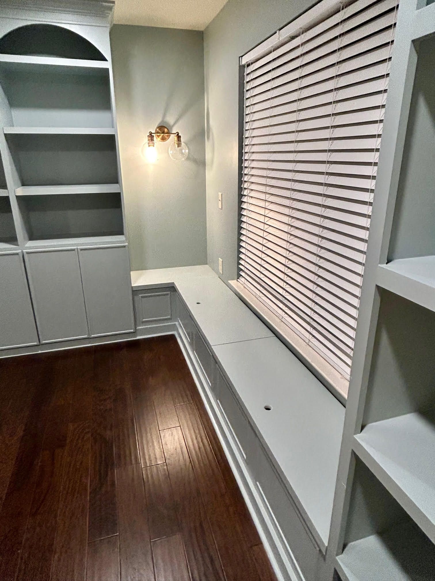 A room with hardwood floors , shelves , and a window with blinds.