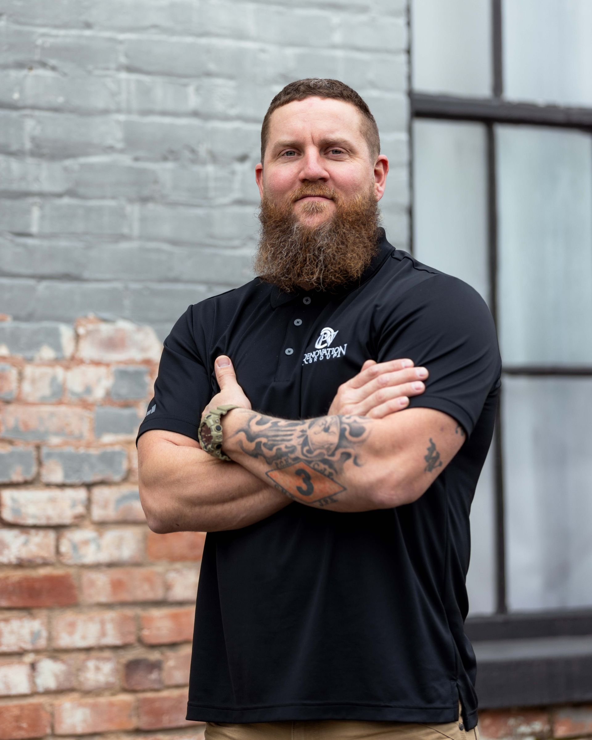 Man with beard, arms crossed, wearing black polo shirt, standing by brick wall, window in background.