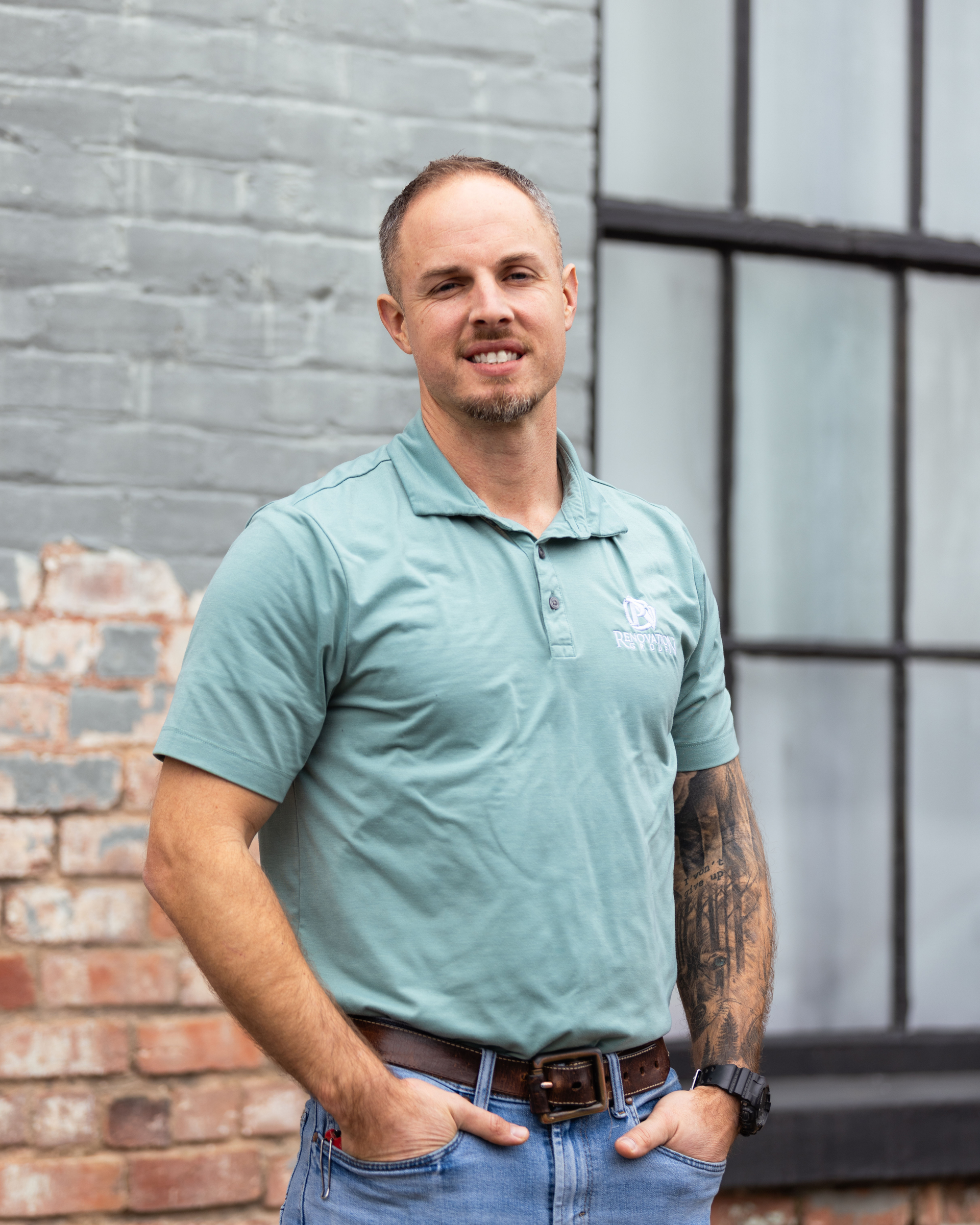 Man in teal polo shirt, hands in pockets, smiling, standing by brick wall and window.