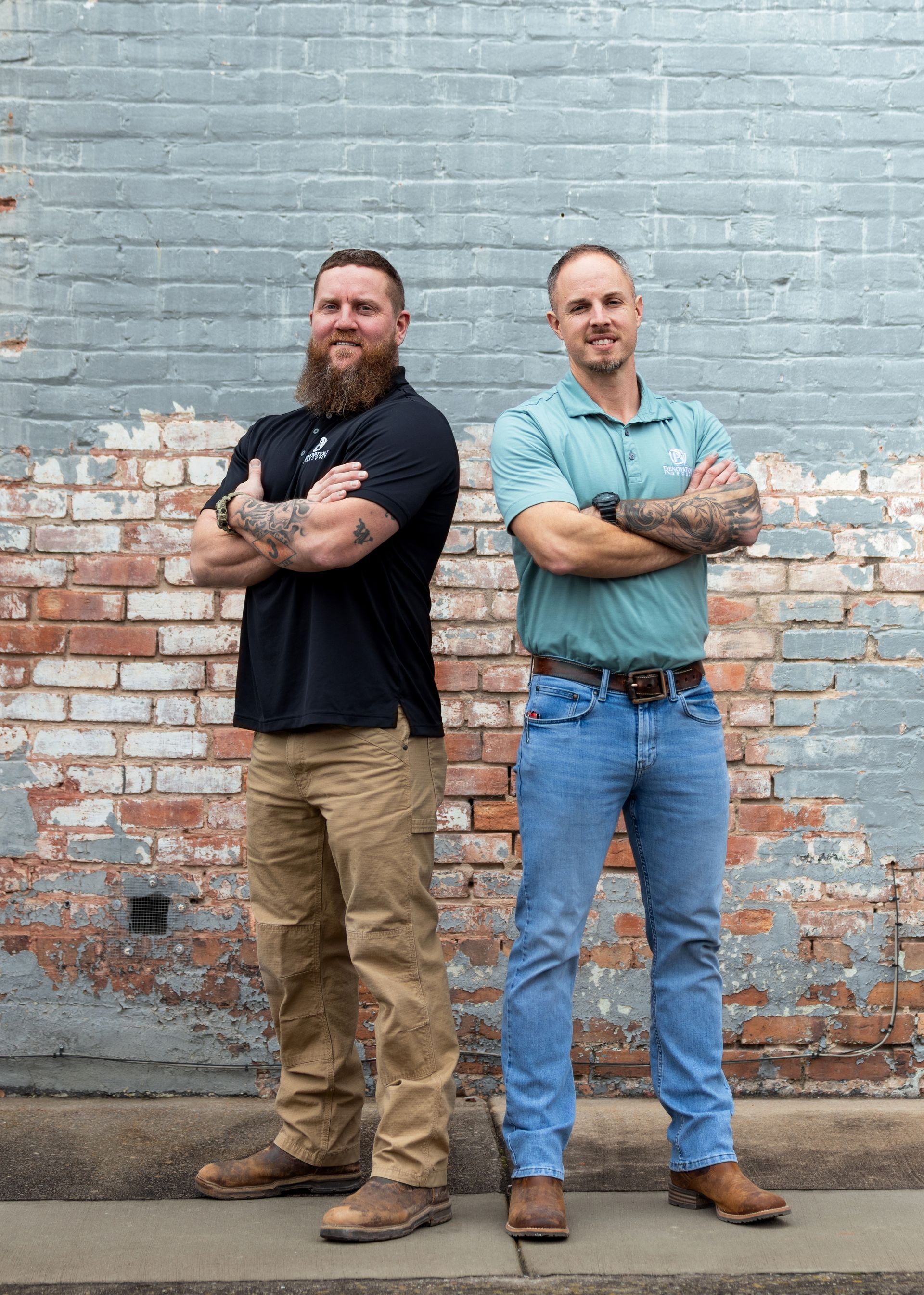 Two men with crossed arms standing in front of a brick wall. One with a beard, black shirt, and khakis, the other in blue jeans and a teal shirt.