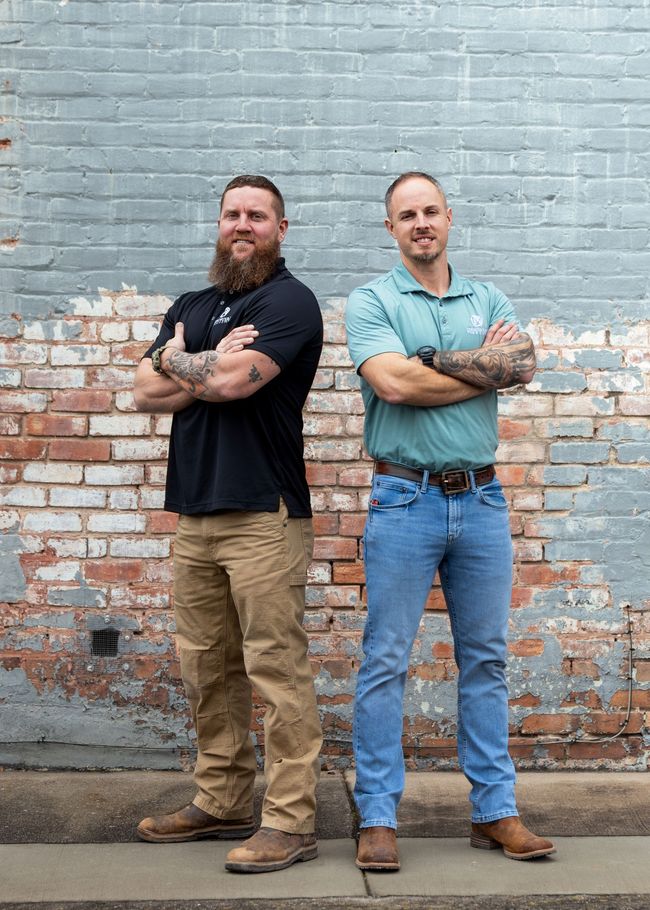 Two men with crossed arms standing in front of a brick wall. One with a beard, black shirt, and khakis, the other in blue jeans and a teal shirt.