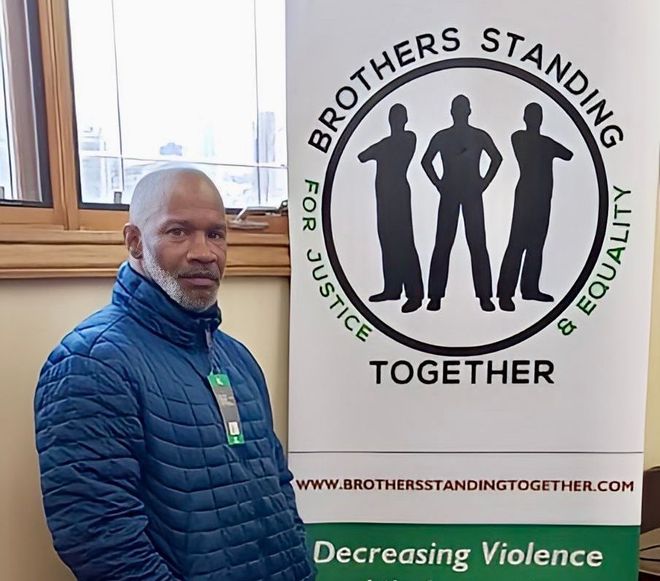 A man in a blue jacket is standing in front of a sign that says brothers standing together