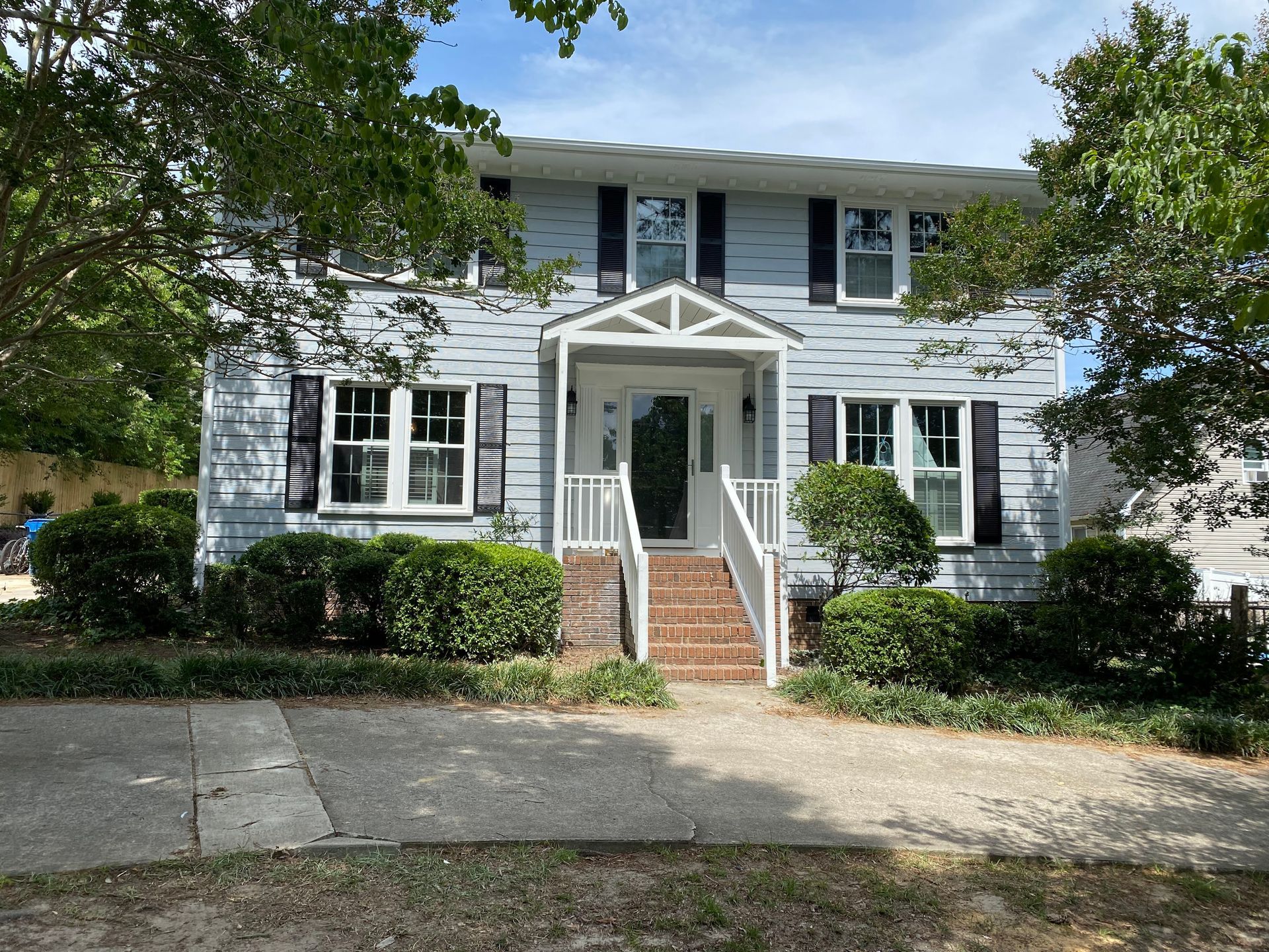 A large house with a porch and stairs is surrounded by trees and bushes.