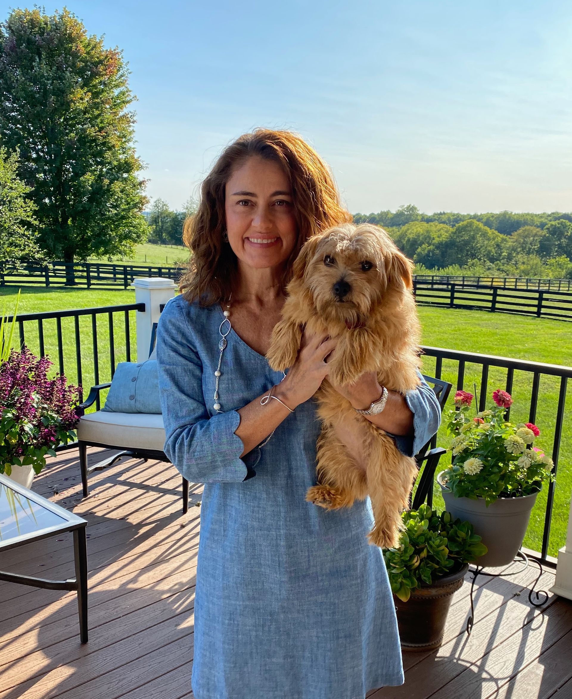 Woman with curly hair holds small, tan dog on a porch overlooking a green field.