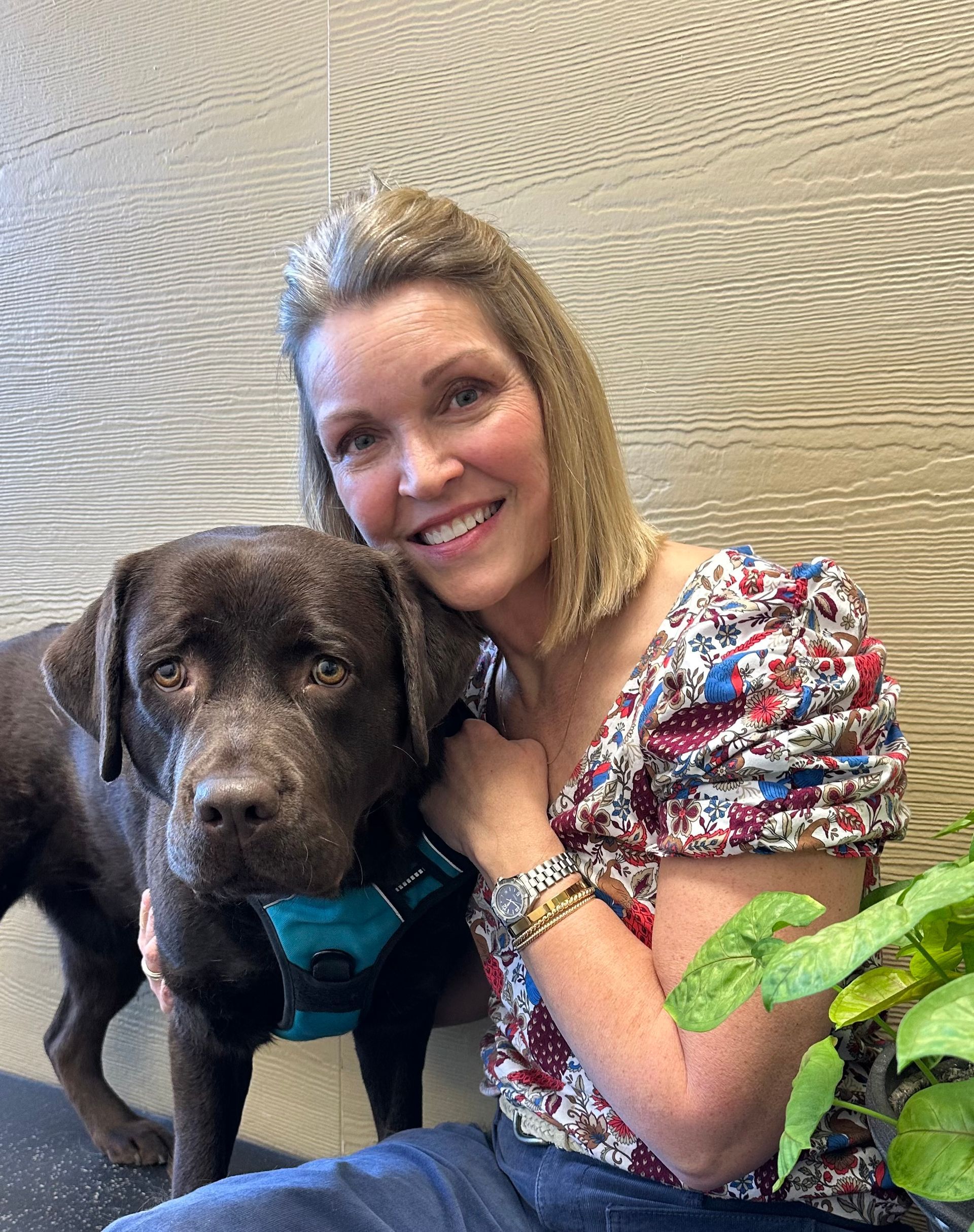 Woman smiling with brown dog, both looking at the camera. They're indoors. The woman has light hair and is wearing a floral top.