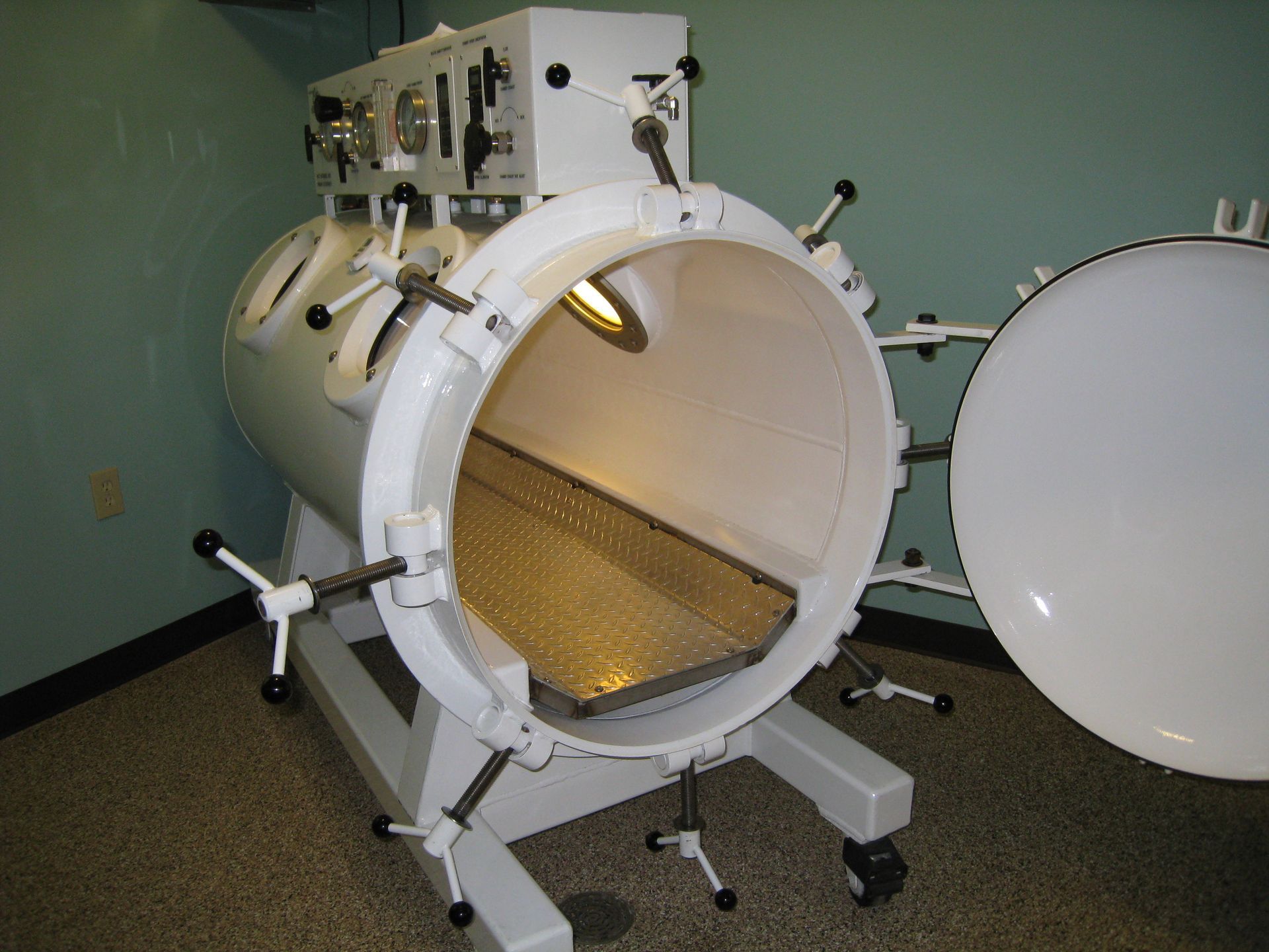 White hyperbaric chamber with open door, stainless steel interior, mounted on a mobile base in a room.