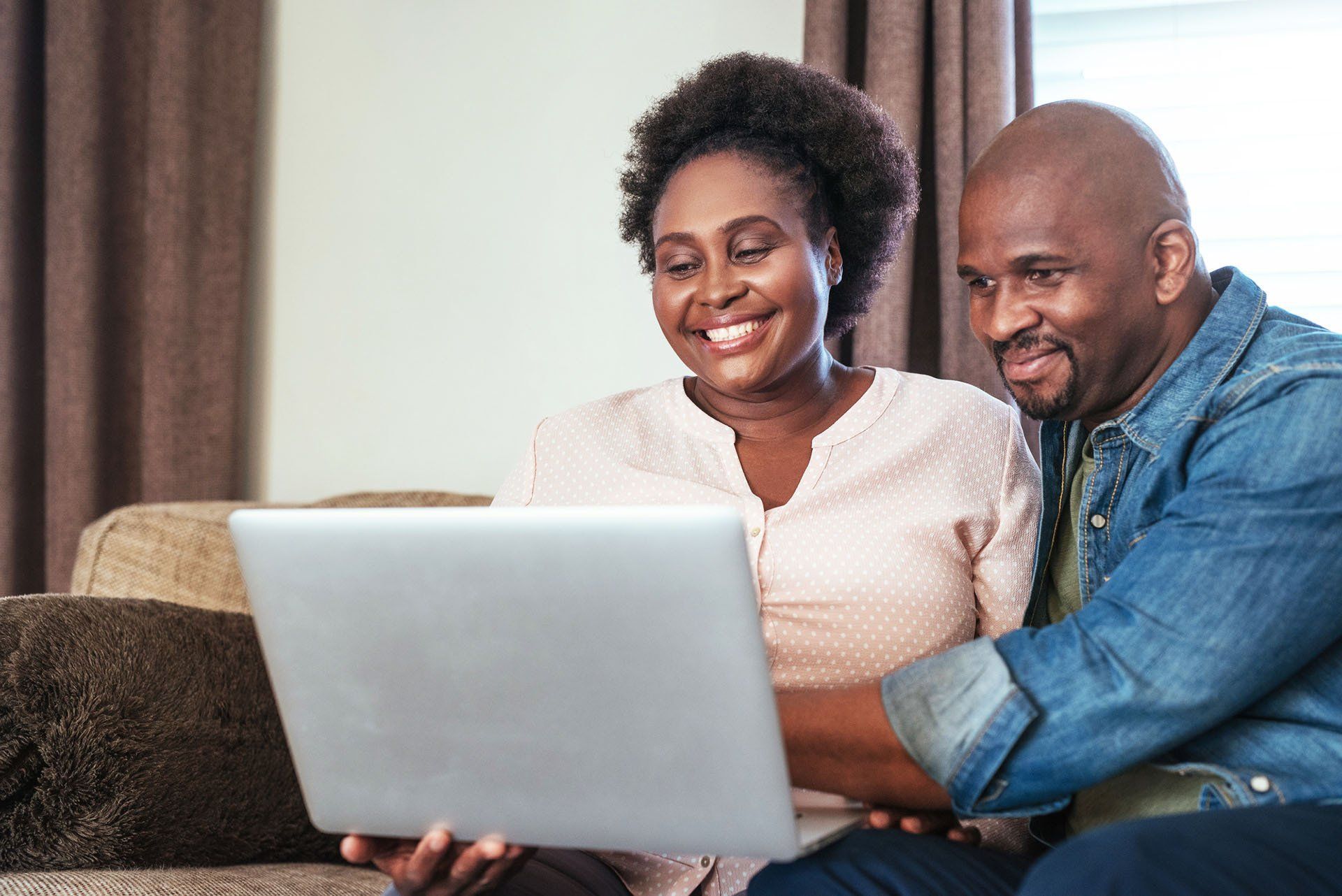 A man and woman sit on a sofa, smiling as they look together at a laptop screen in a brightly lit room.