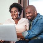 Two people smiling while looking at a laptop screen together in a cozy indoor setting.
