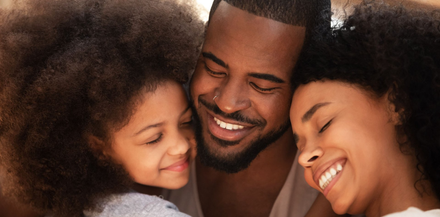 A parent smiles while being hugged by two children, all with their eyes closed in a warm, close-up family moment.