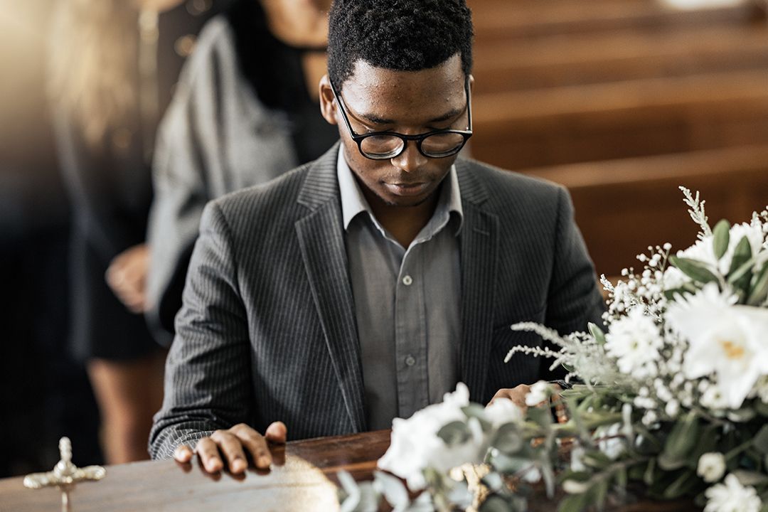 A person wearing a suit and glasses stands somberly by a casket adorned with white flowers at a funeral.