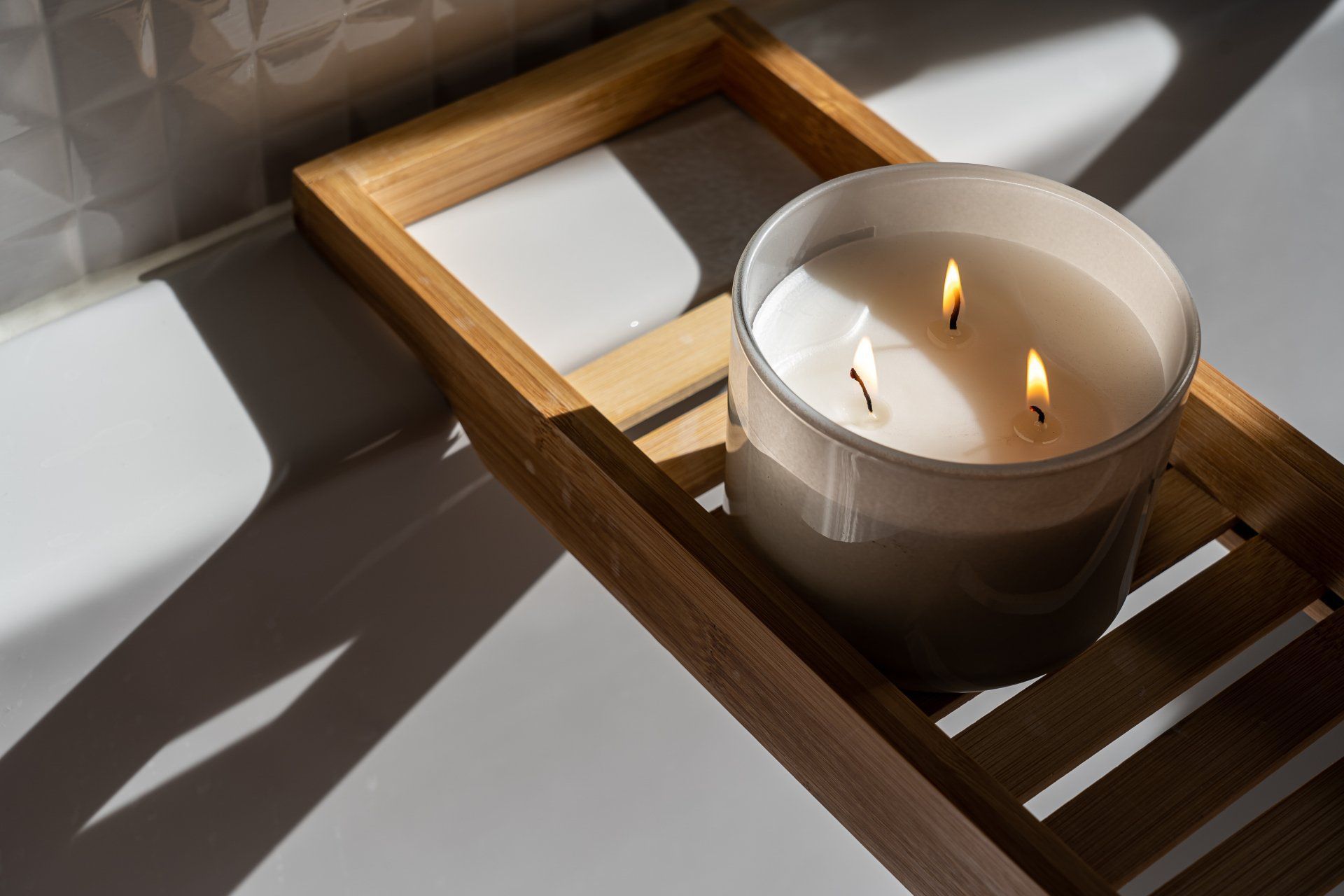 Candle with three lit wicks in a glass jar sits on a wooden bath tray, with sunlight casting shadows.